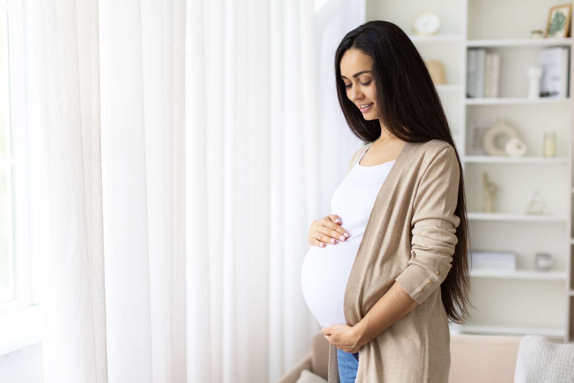 Pregnant young woman peacefully looking at belly while standing by the window