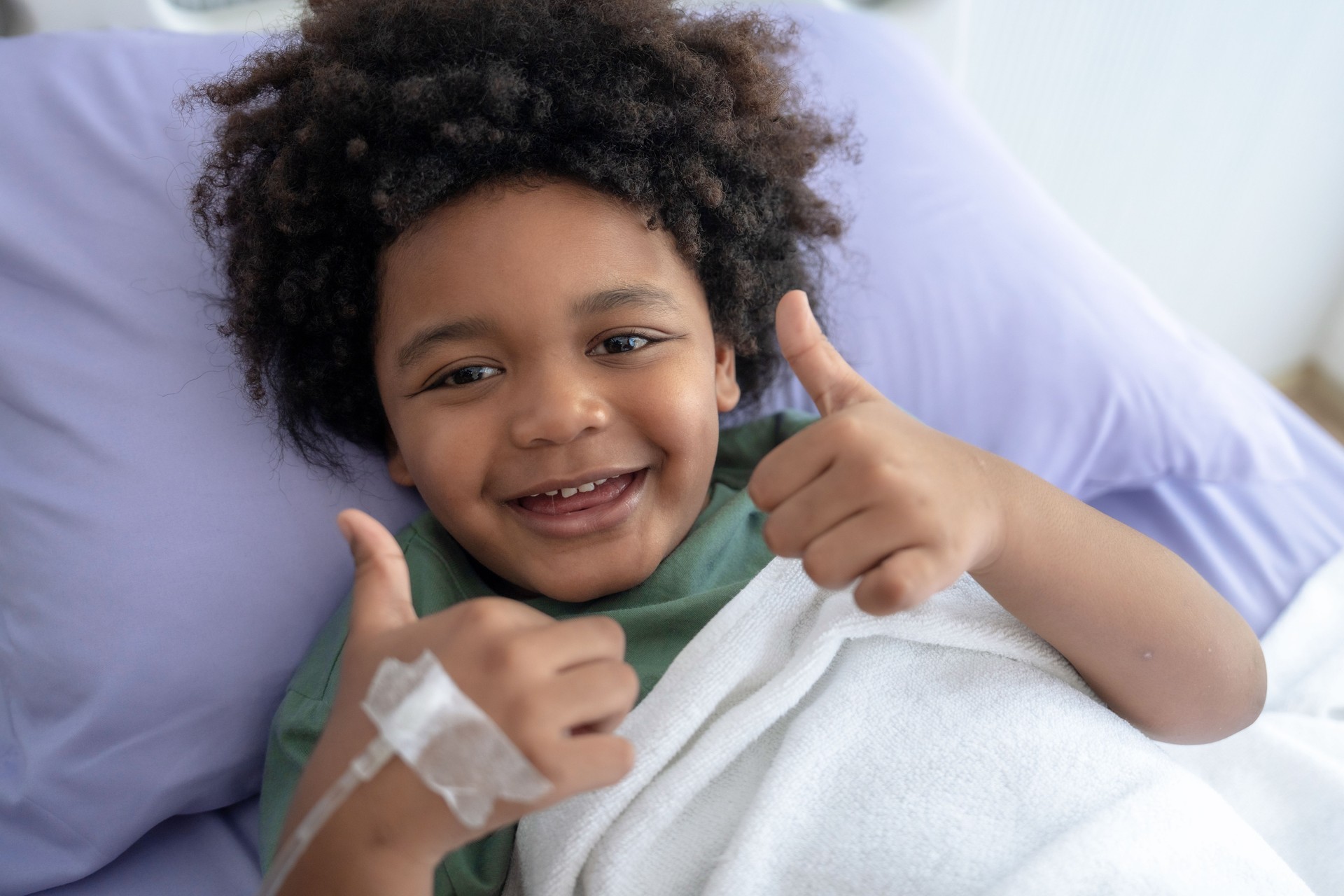 A little boy lies on the hospital bed and smiles back at the camera.